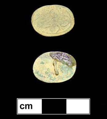 Flat copper alloy button with a heavily worn floral motif. There appears to be one four-petaled flower with the petals on the long axis of the oval shape larger than the petals on the short axis. Discoloration on the back is the artifact label.